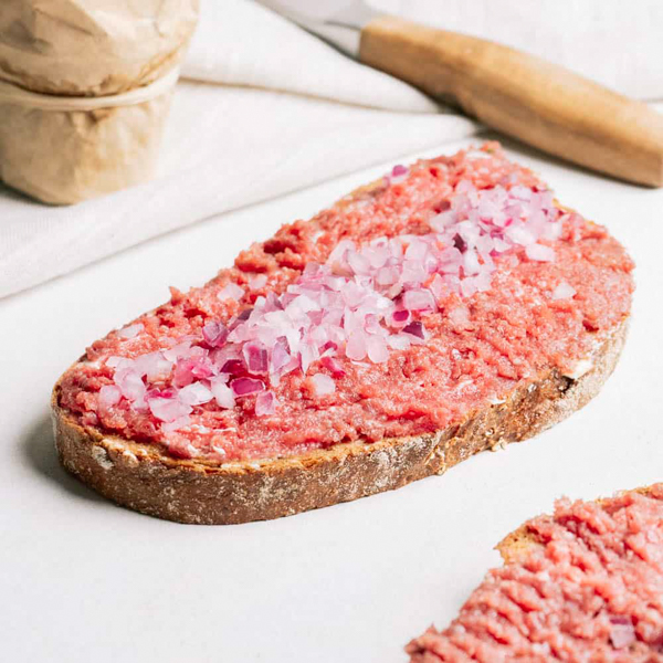 Genussmacher ﻿Eine dicke Scheibe Krustico belegt mit Rindermettwurst mit Rapsöl nach Art einer Zwiebelmettwurst und roten Zwiebeln.